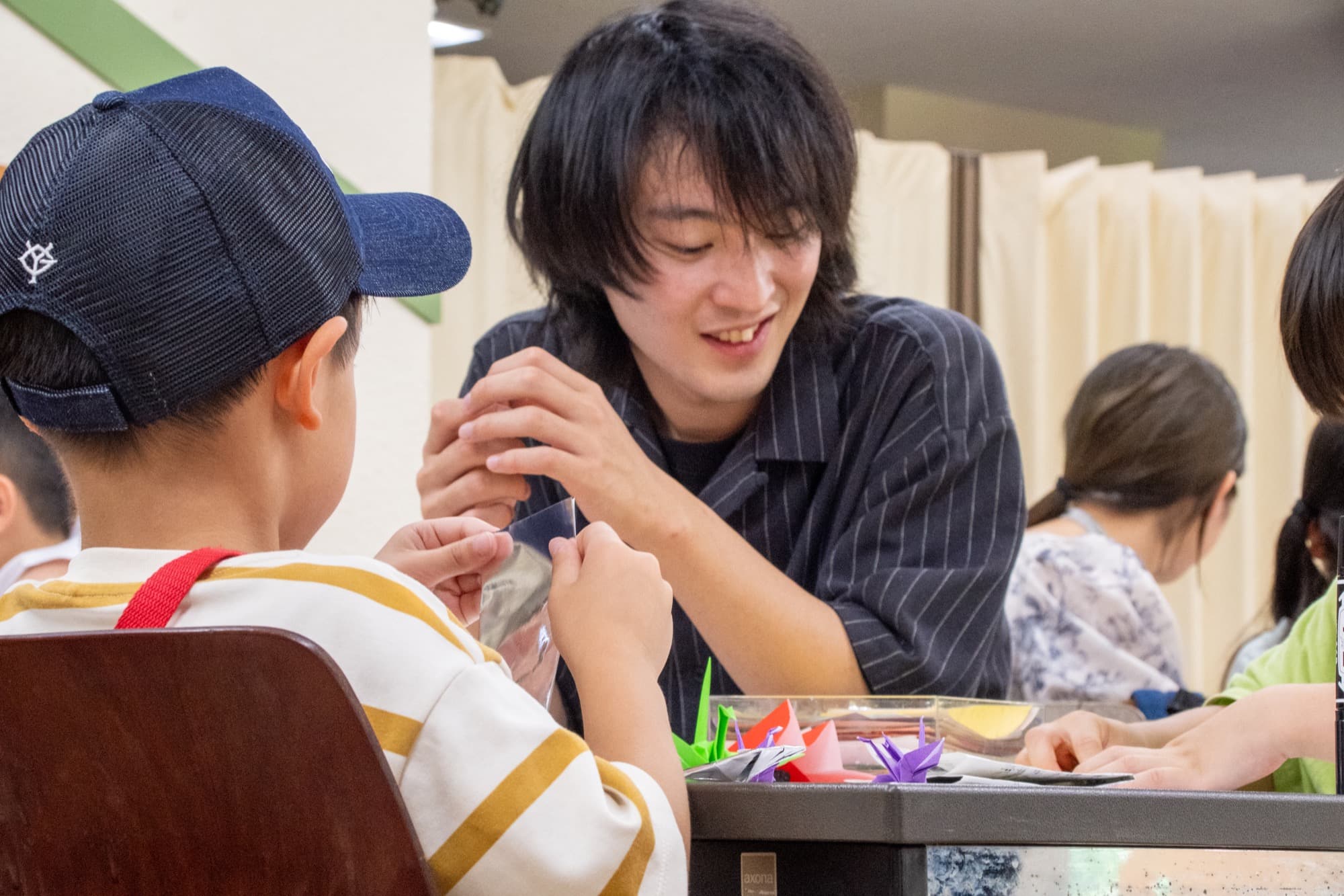折り紙を一緒に折る大学生と子ども(子どもは後ろ姿)