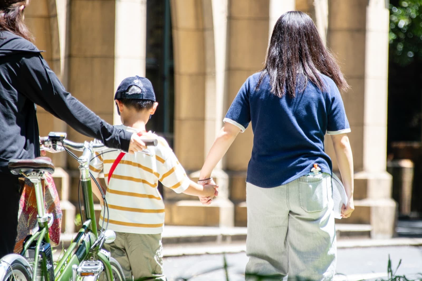 自転車を引く大学生と手をつないでキャンパスを歩く子ども(後ろ姿)