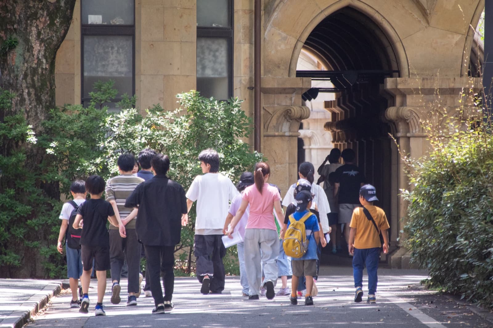 キャンパスの建物へ向かって歩く子どもと大学生の一行(後ろ姿)